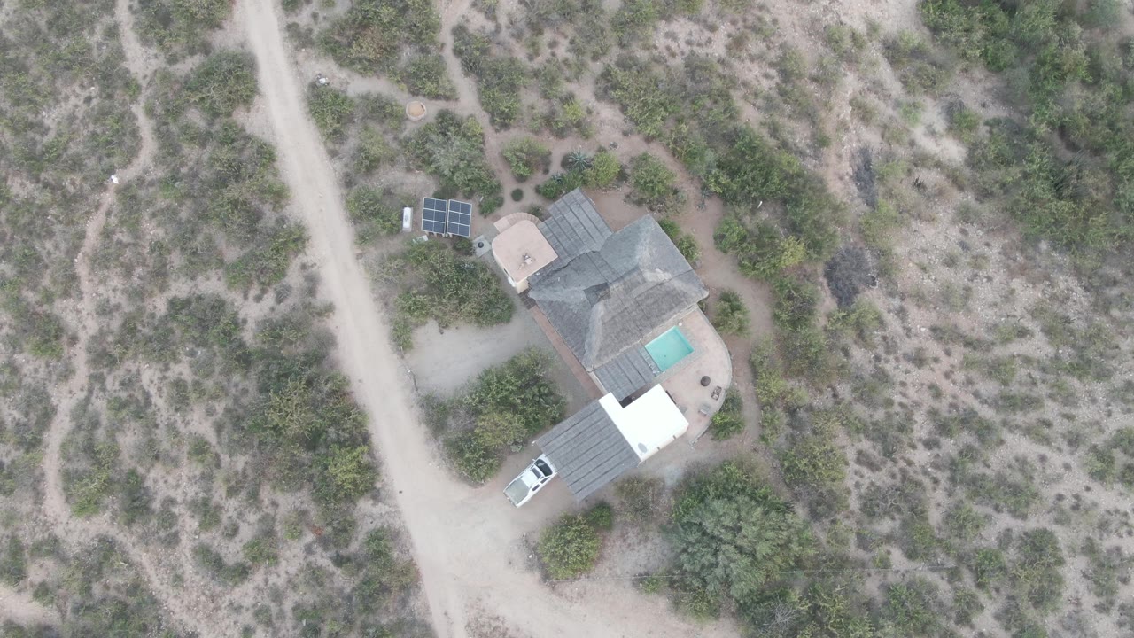 Overhead aerial frame in Cabo Pulmo, Baja California Sur, Mexico showing a pale dirt road, an isolated house, a turquoise pool, solar panels, scrub vegetation, and no visible people.
