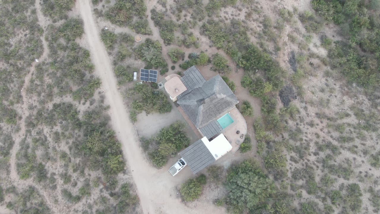 Aerial view in Cabo Pulmo with the white car slightly farther along the dirt road beside a small house, pool, solar panels, dry scrubland, and no visible people.