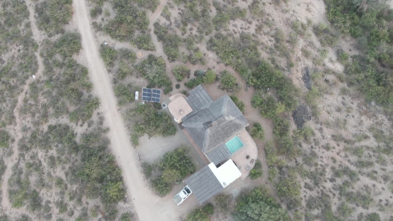 High drone frame over Cabo Pulmo terrain showing the white car on the dirt road, the isolated house and pool below it, solar panels nearby, and no visible people.