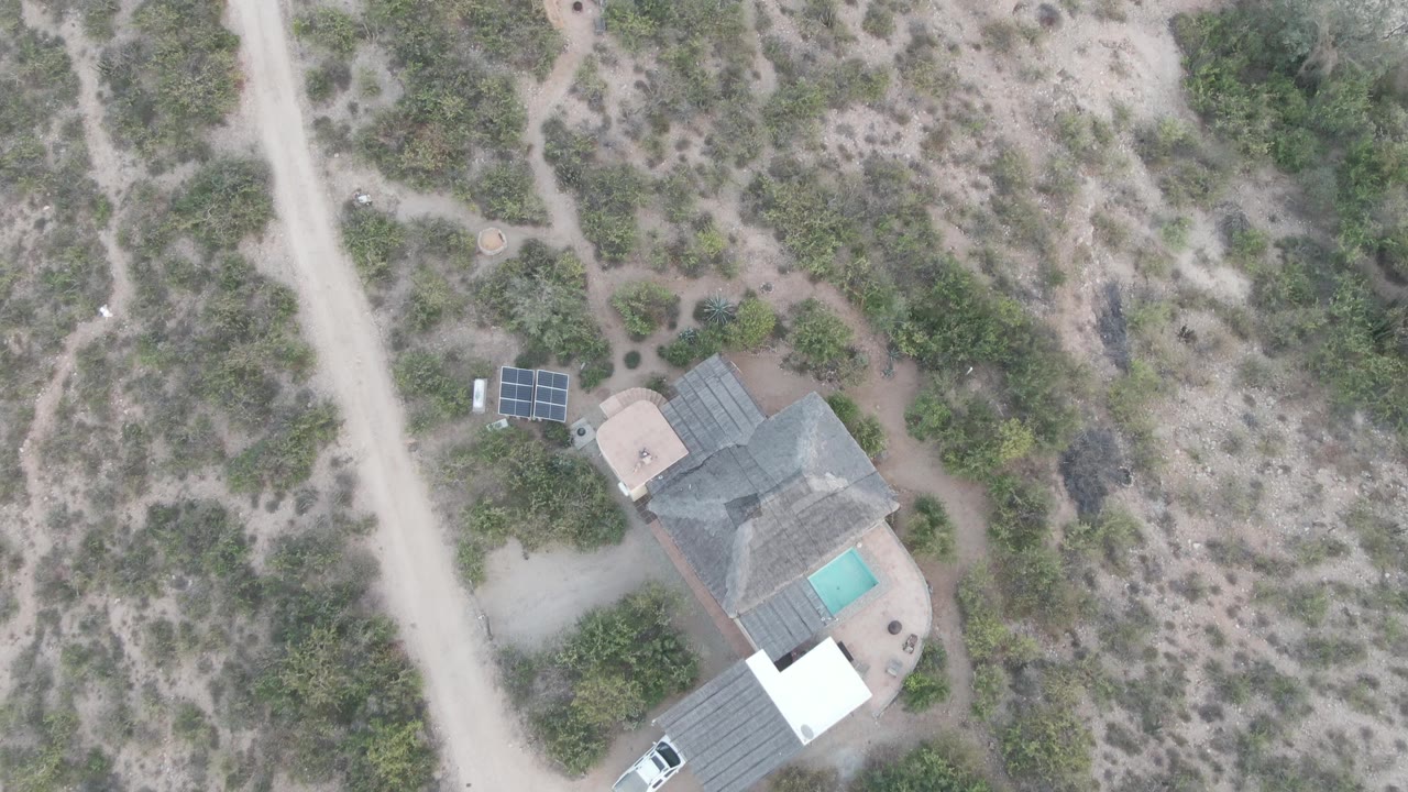 Aerial frame of a dirt road, scrub vegetation, isolated house, pool, and white car.