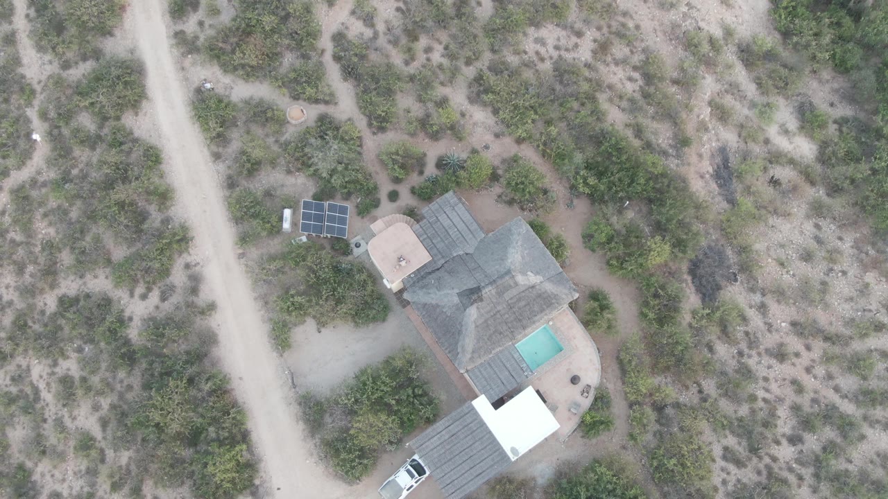 Overhead frame in Cabo Pulmo showing a white car near a pale dirt road, a desert home with a small pool and solar panels, scrubby dry terrain, and no visible people.