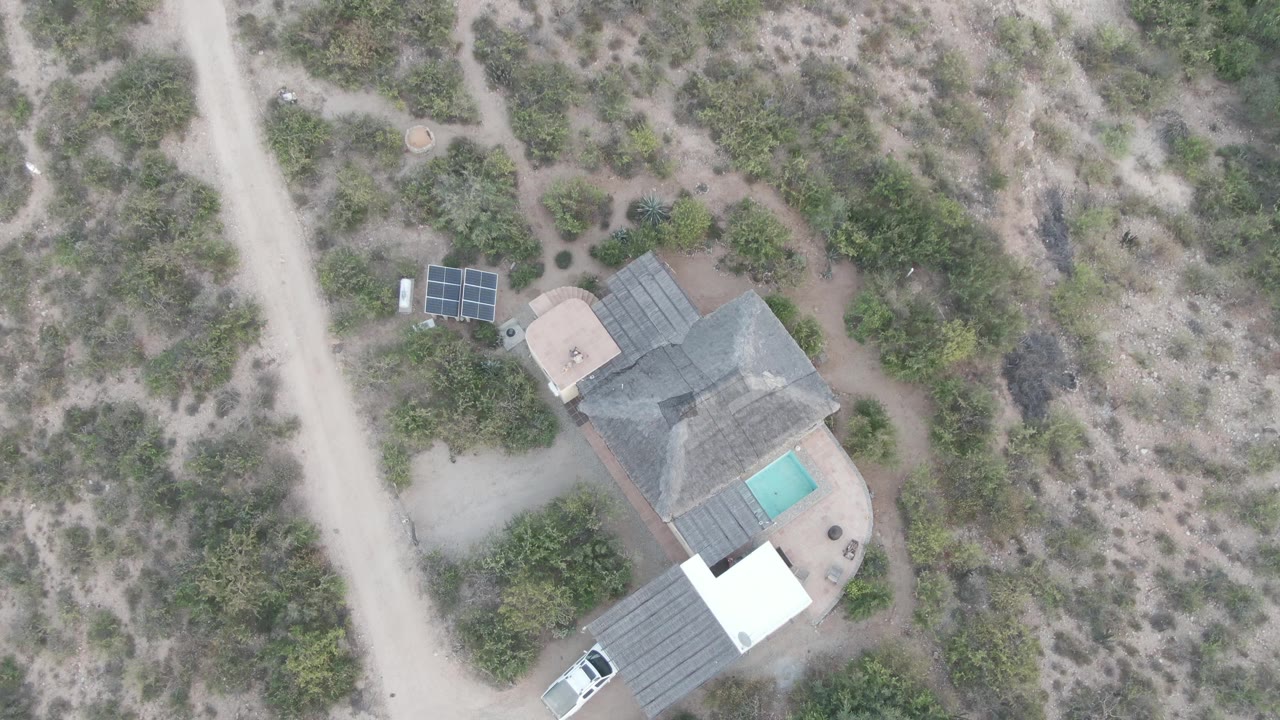 Aerial frame in Cabo Pulmo with the white car slightly farther along the dirt road near the lower edge, a roofed house, solar panels, a turquoise pool, dry shrubs, and no visible people.