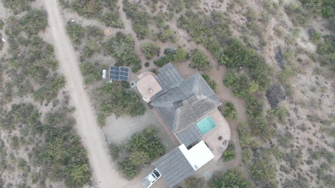 Drone frame in Cabo Pulmo showing the house and pool centered lower in frame, solar panels near the road, the white car near the bottom edge, surrounding desert brush, and no visible people.