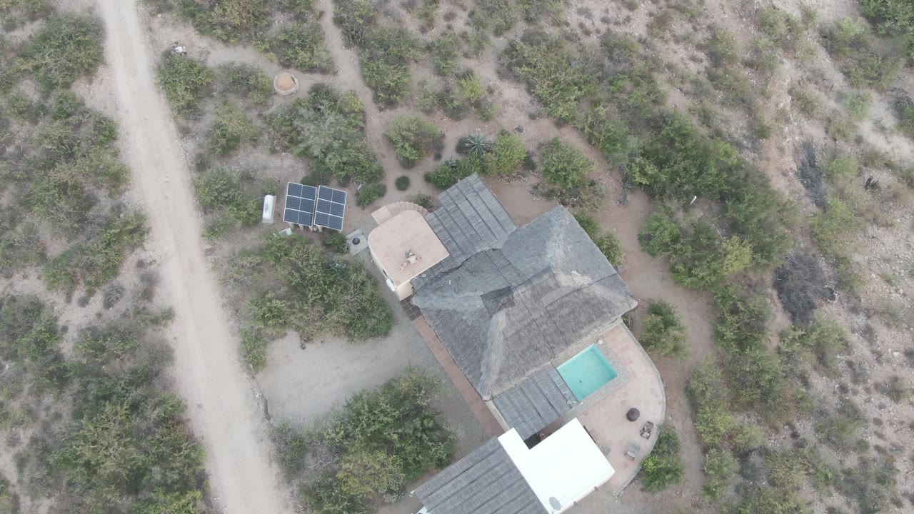 Overhead aerial frame in Cabo Pulmo, Baja California Sur showing a gray-roofed desert house with a turquoise pool, solar panels near the road, dry scrub, and no visible people.
