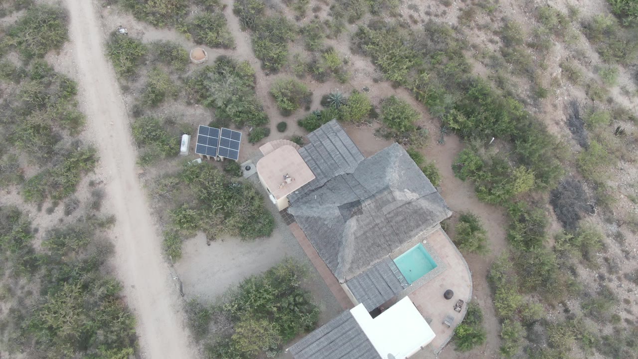 Aerial view of the same desert house with solar panels near a dirt road and a pool beside the roof.