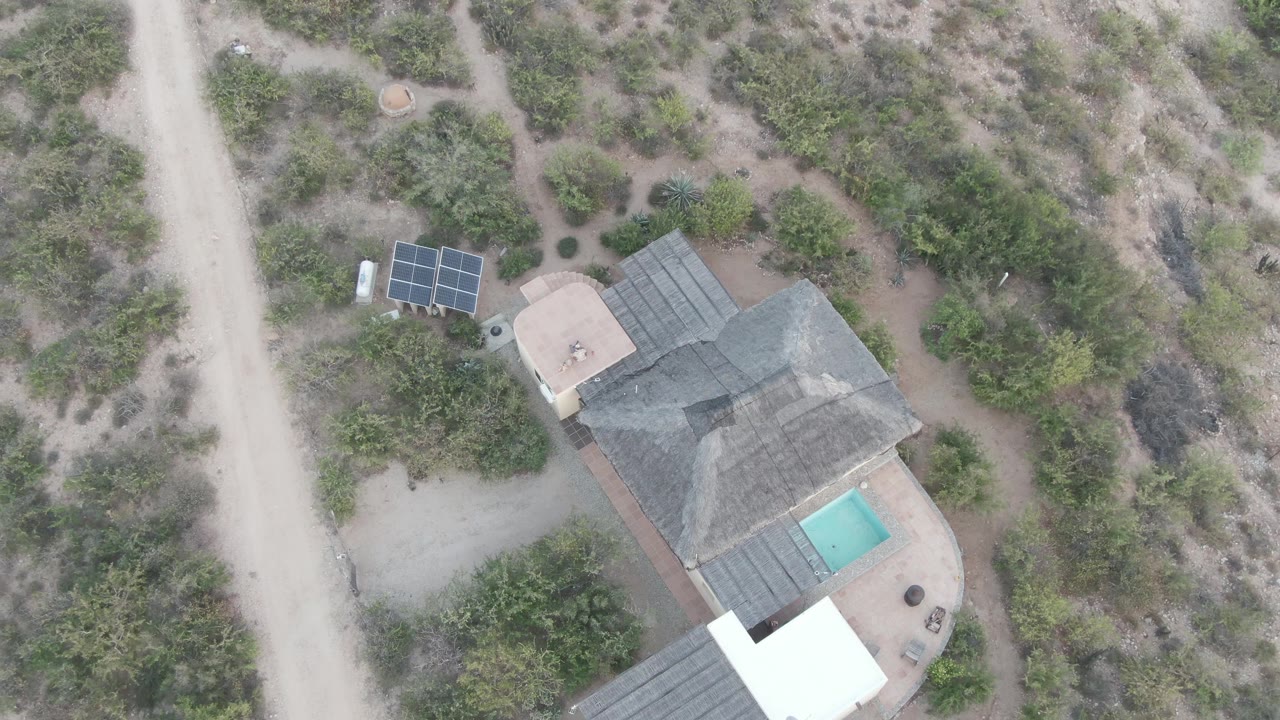 Overhead Cabo Pulmo frame showing the desert property slightly farther along the pass, with the white-roofed structure more prominent at the bottom edge, solar panels above the house, and no visible people.