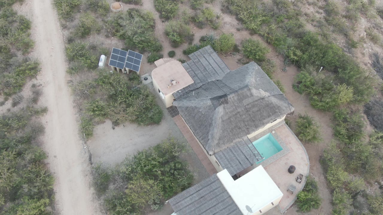 Overhead frame of a Cabo Pulmo desert house with solar panels at upper left, a small pool beside the roof, a carport at bottom edge, dirt roads, shrubs, and no visible people.