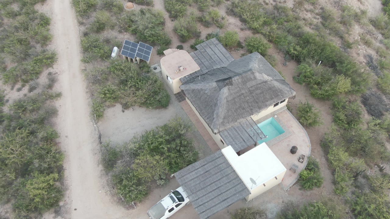 Overhead view in Cabo Pulmo with the white SUV more exposed near the lower carport, the pool to the right of the house, solar panels at upper left, dirt tracks, shrubs, and no visible people.