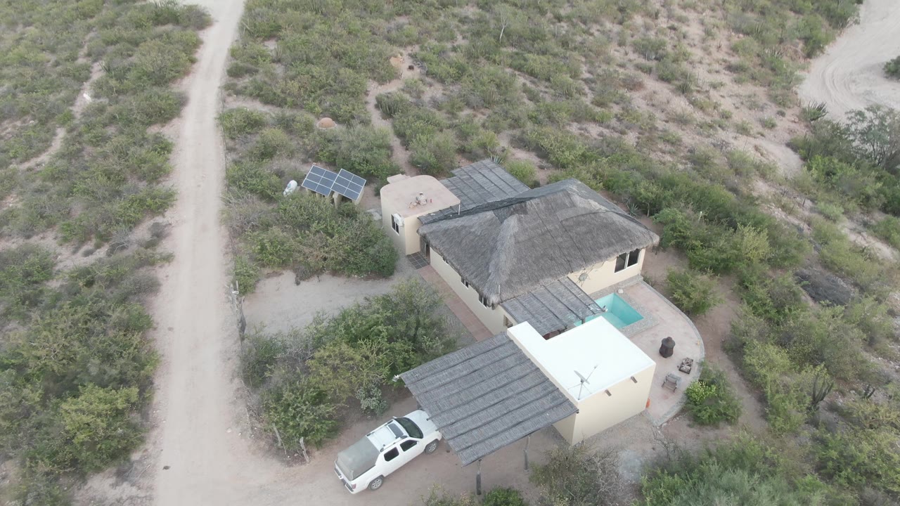 Aerial frame over a rural house in Cabo Pulmo, Baja California Sur, with a dirt road, white pickup, covered patio, solar panels, dry vegetation, and no visible people.