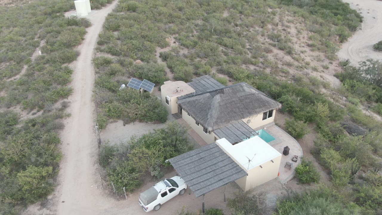 Elevated Cabo Pulmo frame showing the pale house, roof shade, nearby solar panels, curving dirt road, scrubby desert ground, and no visible people.