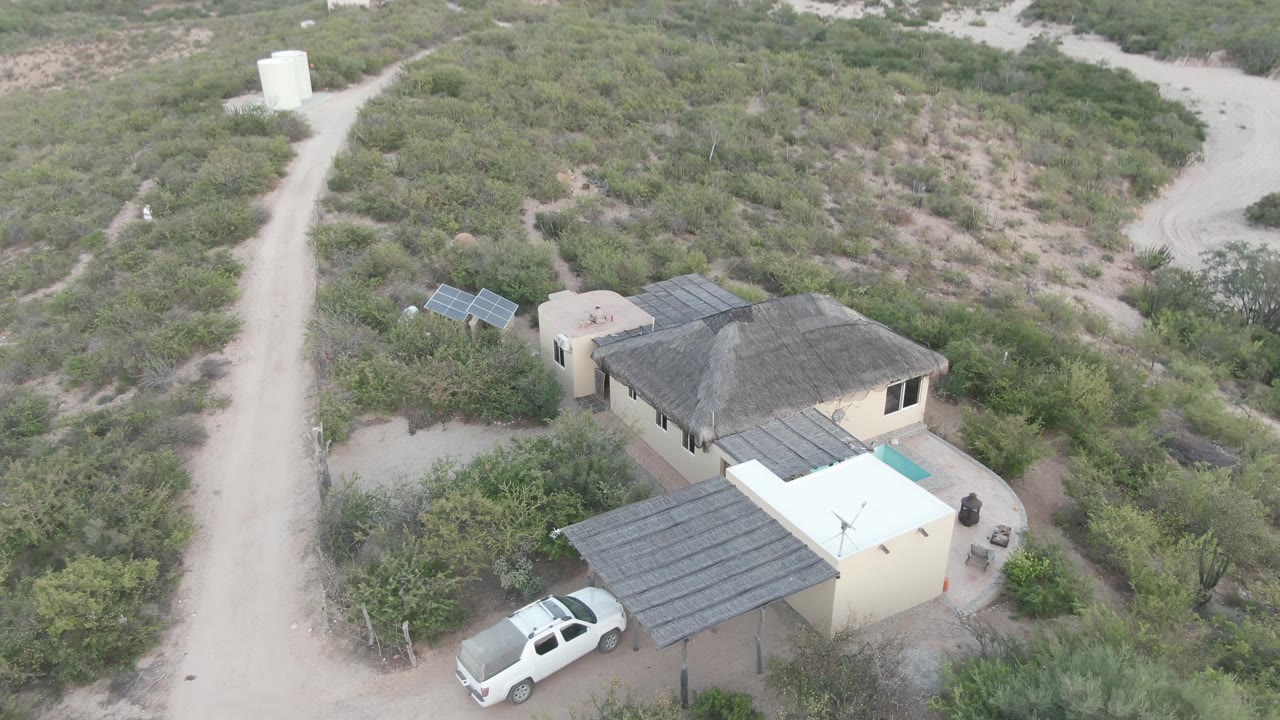 Aerial Cabo Pulmo view with the house and white truck lower in frame, a pale dirt road running past dense desert scrub, and no visible people.