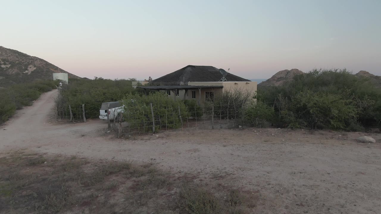 Early frame of a rural house in Cabo Pulmo, Baja California Sur, with a dirt road on the left, fencing and scrub in front, rocky hills behind, and no visible people.