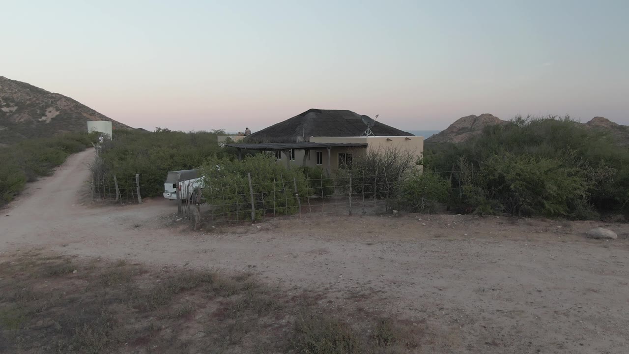 A rural Cabo Pulmo house sits slightly right of the dirt road, with a white vehicle near the fence line, dense brush around the property, rocky hills in back, and no visible people.