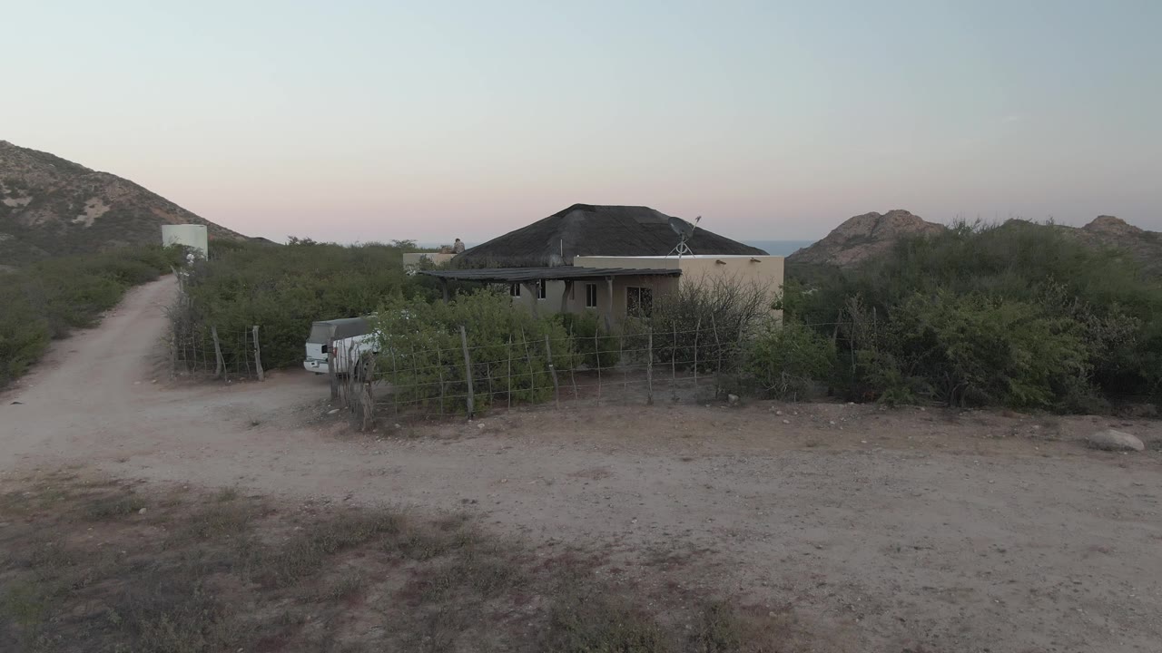 Mid-clip view of the Cabo Pulmo rural property, with the dirt road leading past scrub vegetation, the house roof centered low in frame, hills beyond, and no visible people.