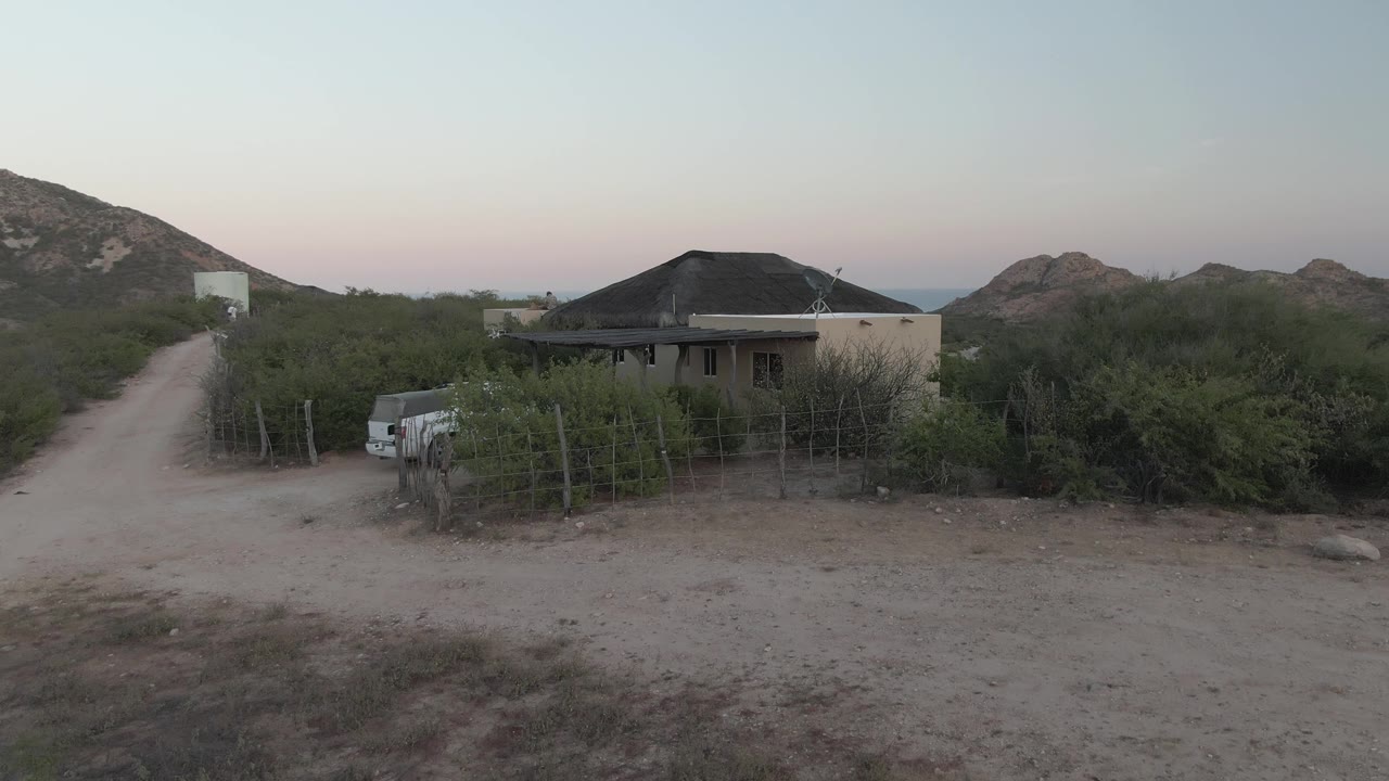 Rural house and dirt track in dry vegetation under a pale dusk sky.
