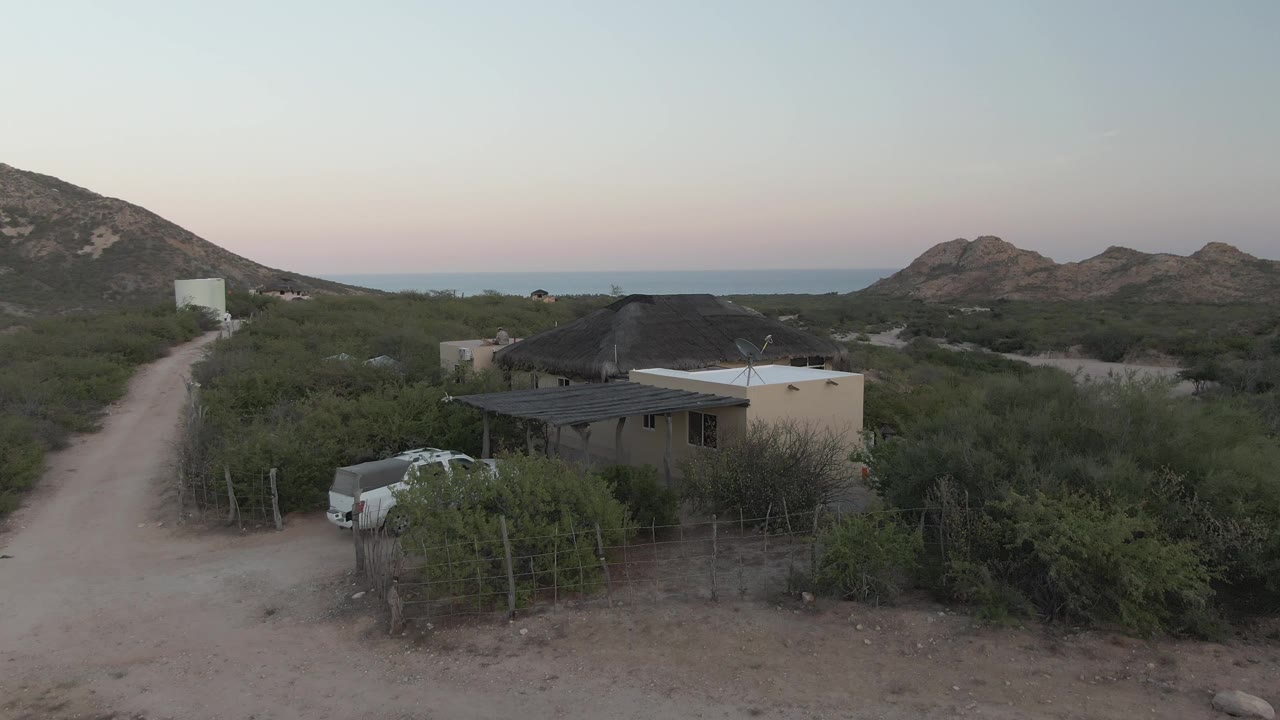 Wide elevated view in Cabo Pulmo, Baja California Sur, showing a white pickup beside a dirt road, a thatched-roof house, scrub brush, rocky hills, ocean horizon, and no visible people.