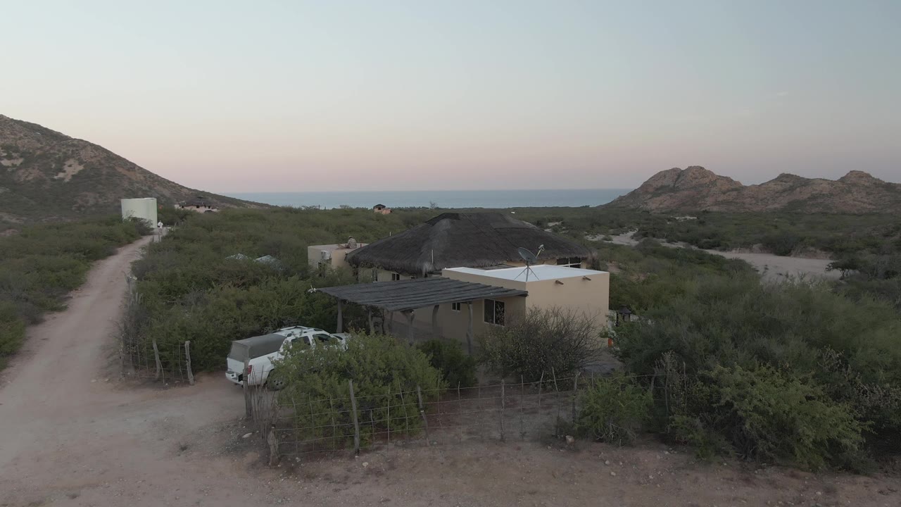 Elevated Cabo Pulmo frame with the dirt road running up the left side, a white pickup near the house, dry green brush, distant rocky hills, ocean band, and no visible people.