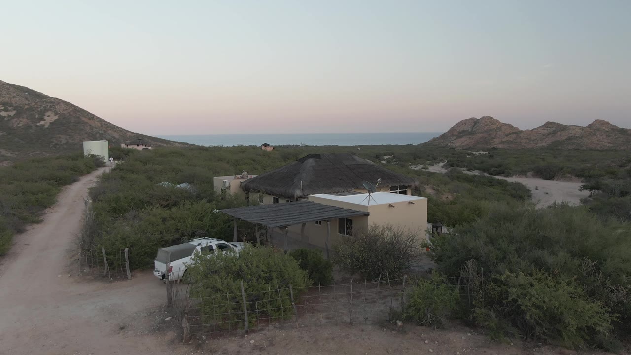 Cabo Pulmo aerial-style view centered on a thatched-roof house and white pickup, with sandy road, fenced brush, rocky hills, ocean horizon, pale evening sky, and no visible people.