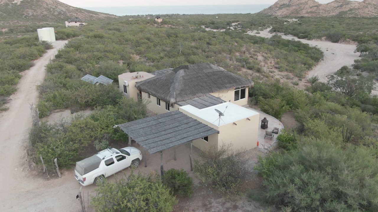 Aerial view in Cabo Pulmo, Baja California Sur, showing a white SUV under a carport beside a pale desert house, scrub vegetation, dirt roads, distant hills, and no visible people.