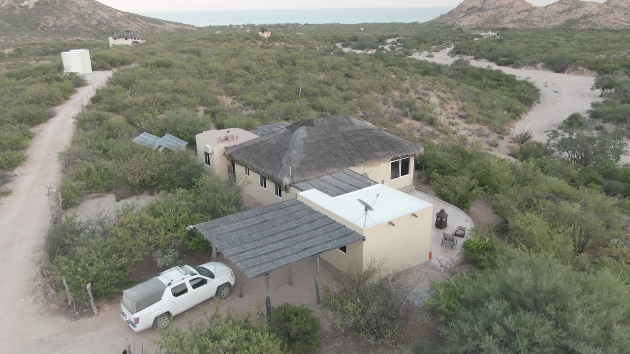 Aerial frame in Cabo Pulmo, Baja California Sur, with the white SUV just beyond the shaded carport, the house roof and patio visible, dirt tracks through shrubs, and no visible people.