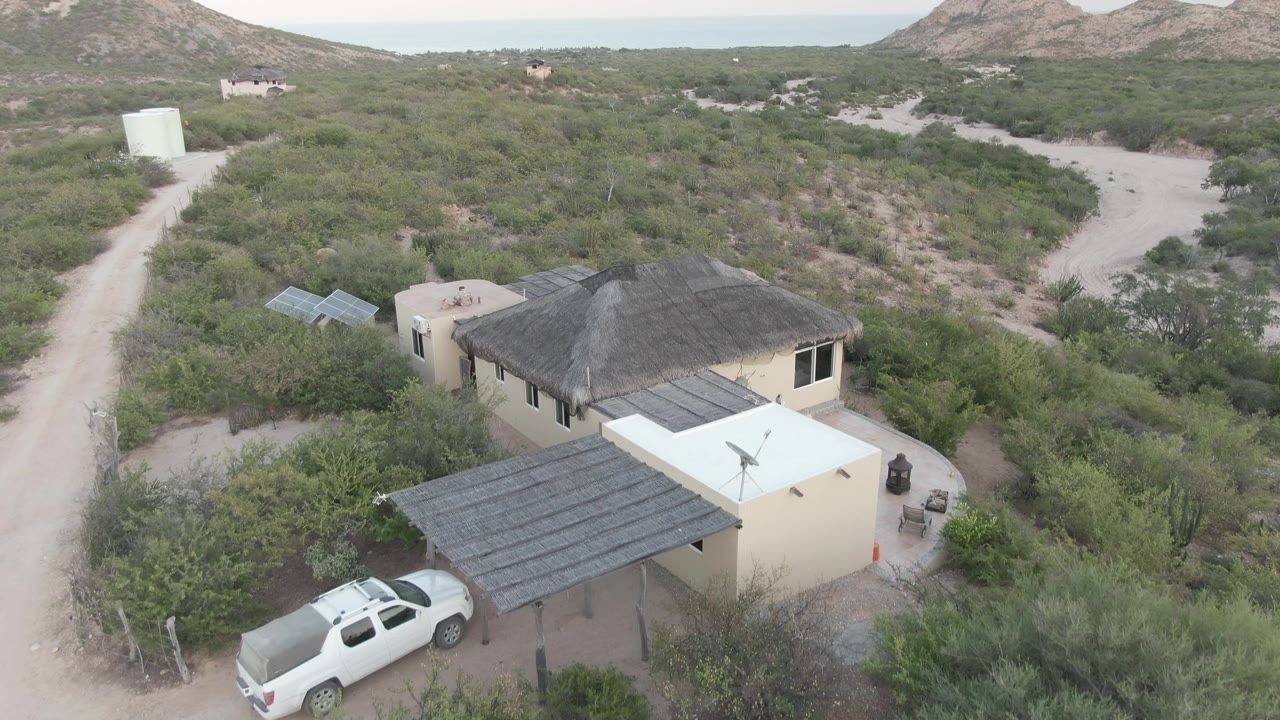 Cabo Pulmo aerial view with the white SUV moving farther from the carport, the pale house and solar panels in the foreground, brushy desert roads behind, and no visible people.