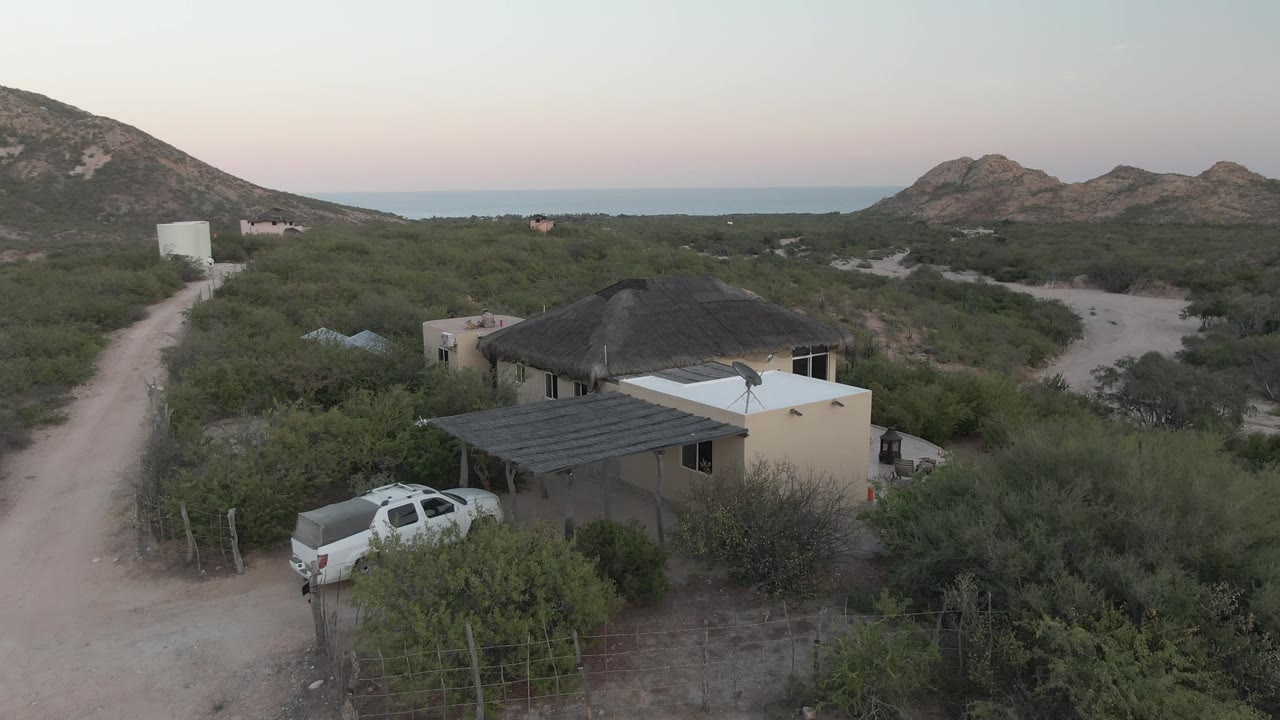 Aerial view in Cabo Pulmo, Baja California Sur, showing a pale desert house, parked white pickup, dirt road, scrub vegetation, rocky hills, distant ocean horizon, and no visible people.