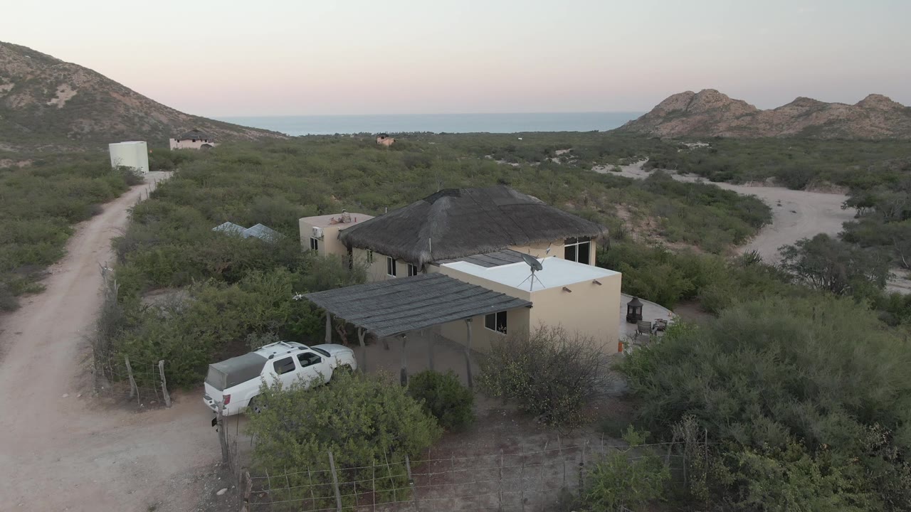 Second aerial frame in Cabo Pulmo shows the drone slightly nearer to the house, with the white pickup at lower left, branching dirt roads through scrub, rocky hills, ocean horizon, and no visible people.