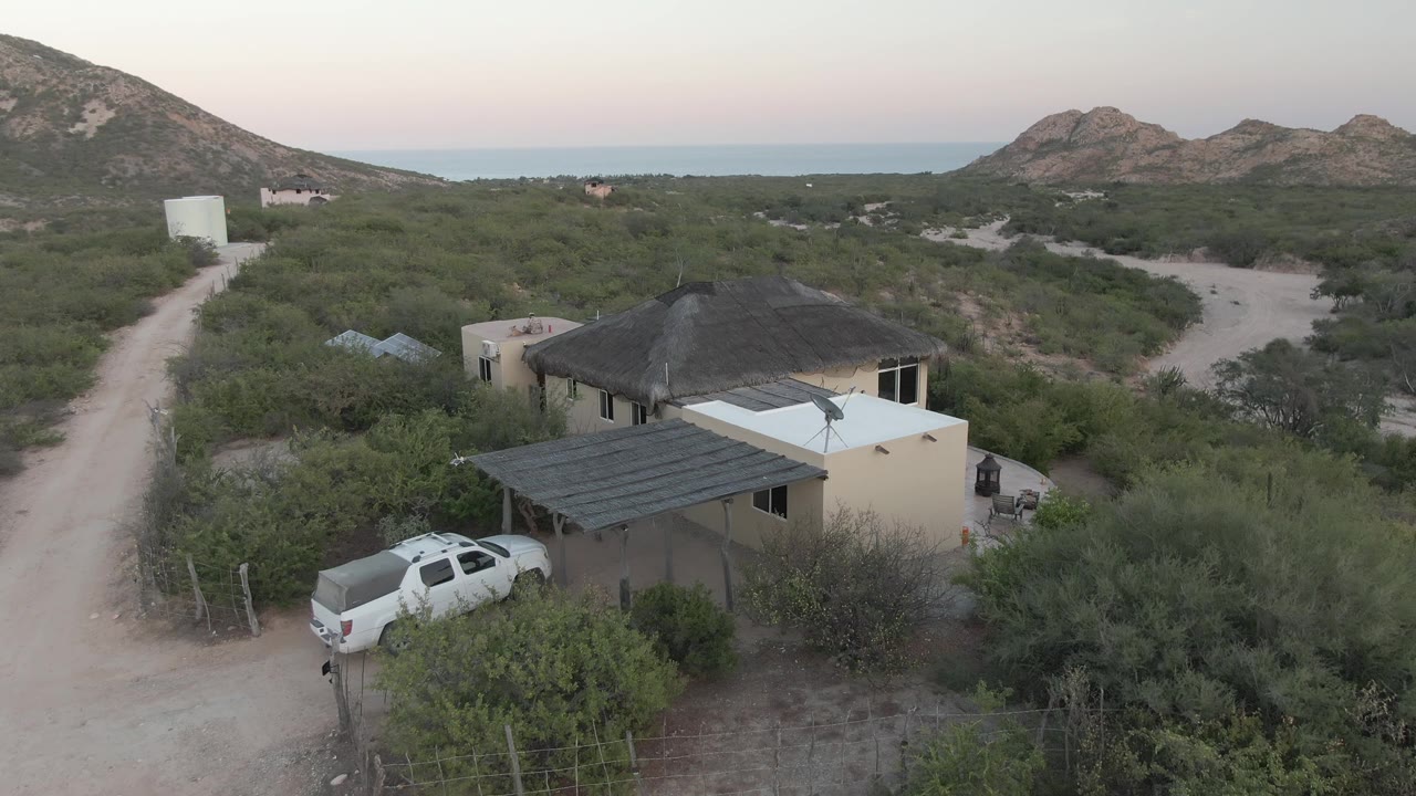 Aerial frame in Cabo Pulmo with the house closer in the lower frame, the white pickup partly screened by brush, dirt tracks running past the property, rocky hills beyond, and no visible people.