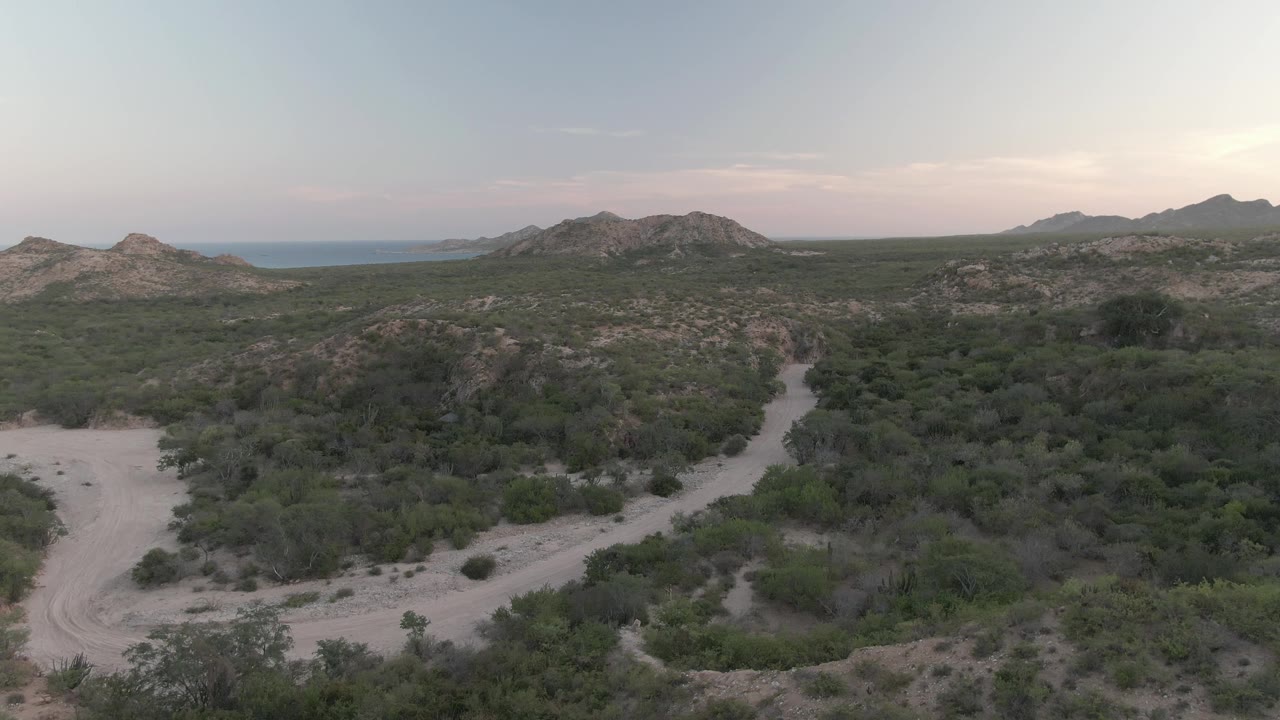 Aerial view over Cabo Pulmo scrubland with a light dirt road cutting through the midground, low hills behind it, distant mountains, and no visible people.