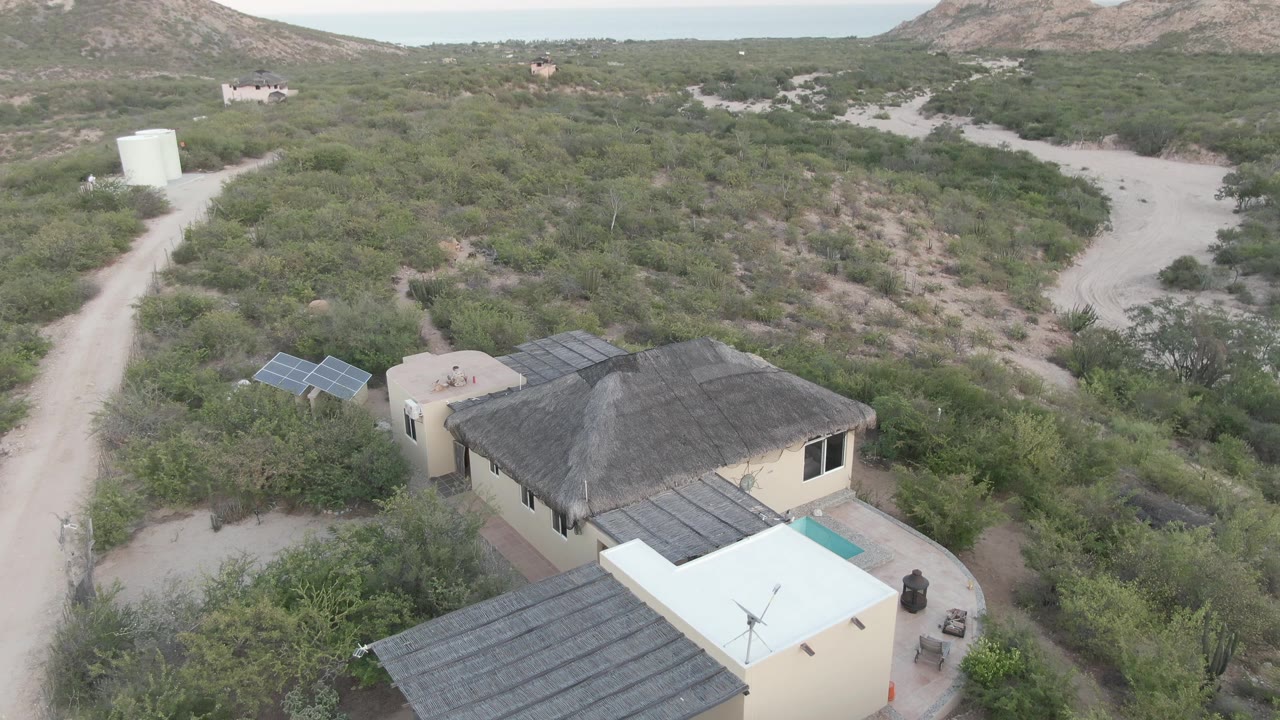 Aerial frame over Cabo Pulmo, Baja California Sur showing a pale rural house, dark roof sections, solar panels, a small turquoise pool, dirt tracks, and scrub-covered hills.