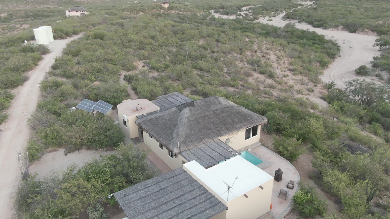 Elevated view of the house roof, nearby solar panels, and a curving dirt road through shrubs.