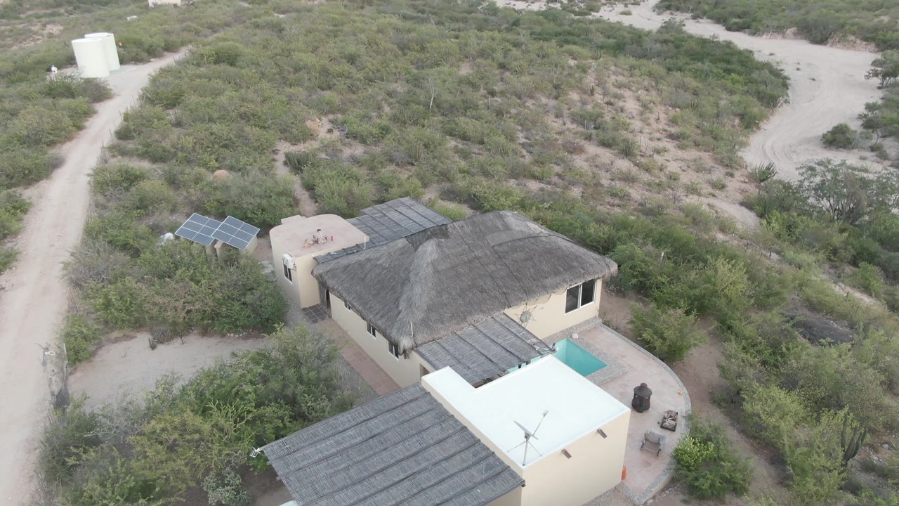 Cabo Pulmo aerial frame showing the house roof, small turquoise pool, solar panel array, branching dirt road, scrub vegetation, and a white cylindrical tank in the distance.