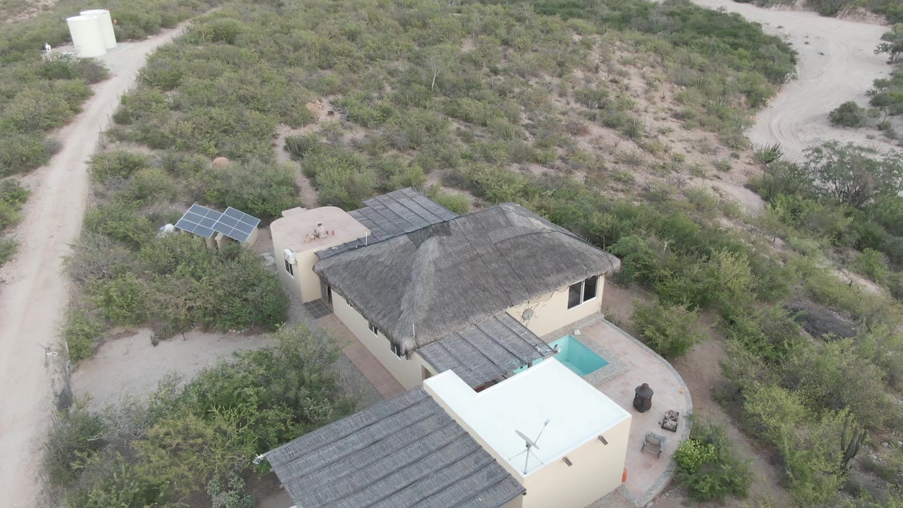 Drone-like overhead view of a house, solar panels, dirt road, and dry hillside vegetation.