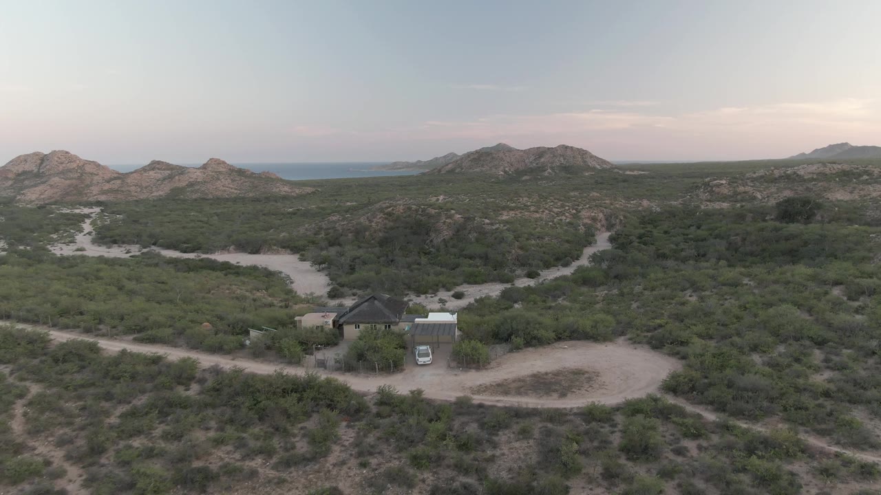 Aerial view over Cabo Pulmo desert scrub with intersecting dirt roads, a small dark-roofed building, a white vehicle near it, rocky hills, distant water, and no visible people.