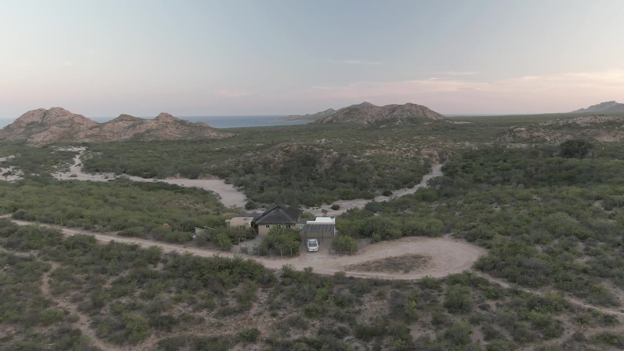 Wide aerial desert landscape with a small building, white vehicle, winding roads, and rocky hills.