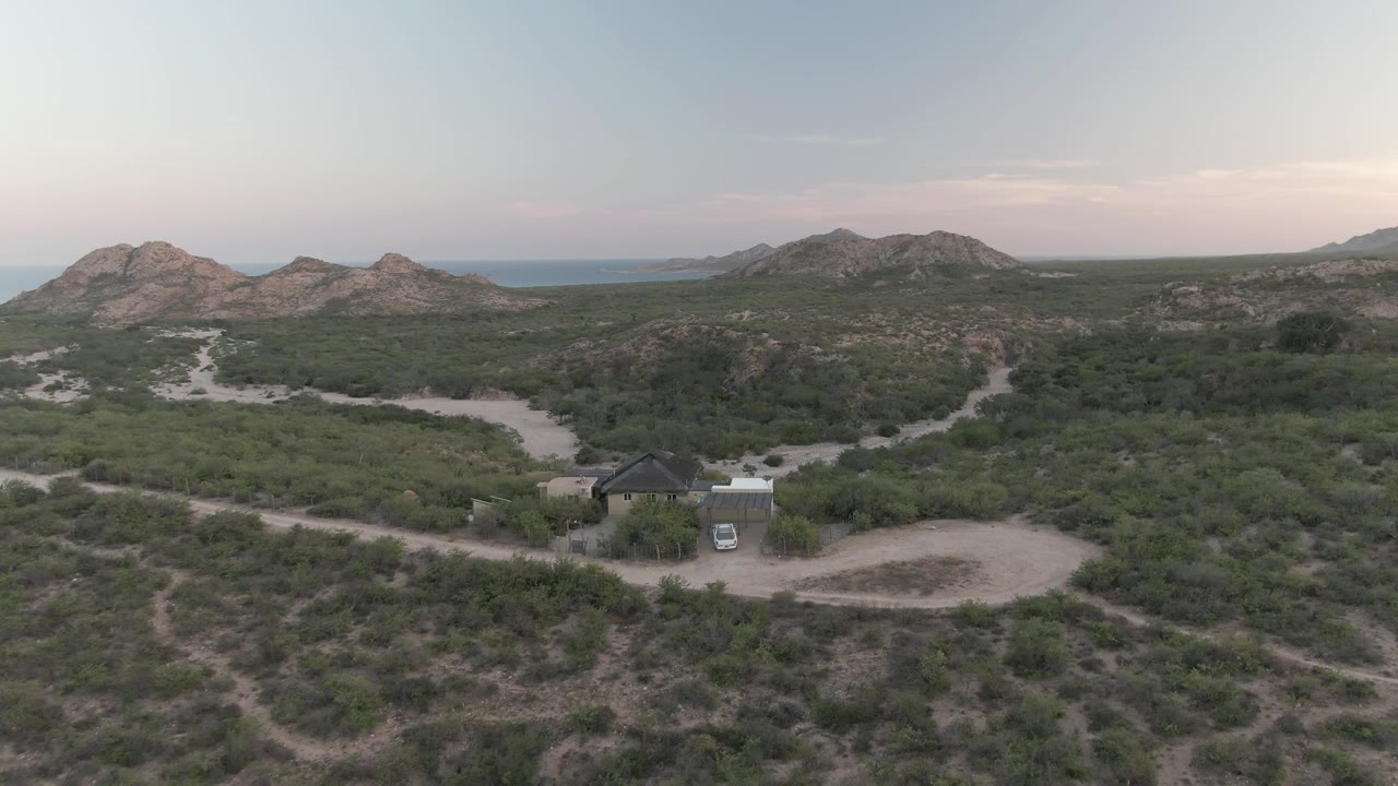 Cabo Pulmo desert aerial with the small building and white vehicle near the lower center, multiple dirt roads crossing the brush, layered rocky hills, distant water, and no visible people.