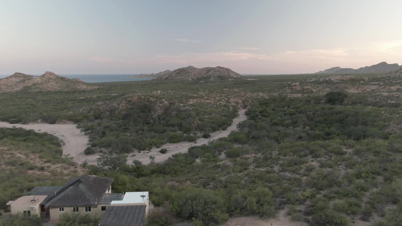 Aerial view over Cabo Pulmo desert scrub with a house and light vehicle at the lower edge, dirt roads through the midground, rocky hills, distant water, and no visible people.