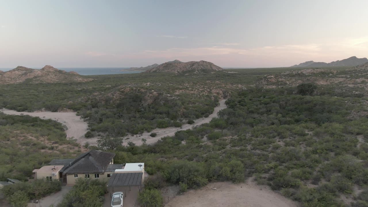 Desert house, parked vehicle, dirt tracks, scrub, hills, and pale sky.