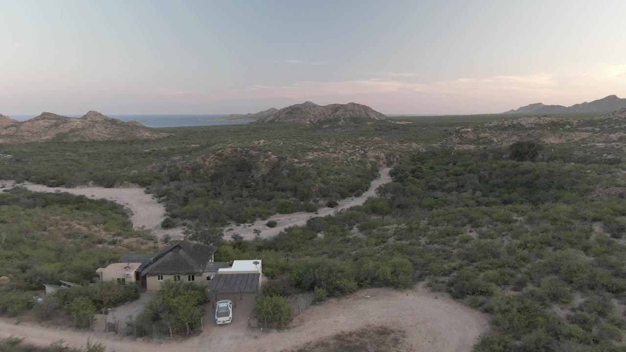 Elevated view in Cabo Pulmo showing the house and vehicle slightly lower in the frame, a sandy loop road in front, dense scrub beyond, rocky hills, distant blue water, and no visible people.