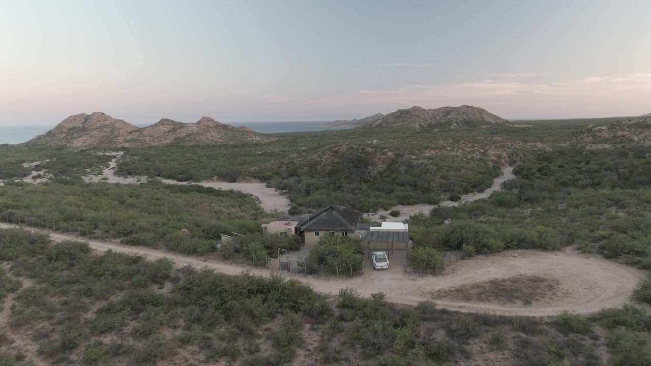 Opening aerial frame over Cabo Pulmo, Baja California Sur, showing a small desert house with a parked car, sandy roads, low bushes, distant rocky hills, and no visible people.