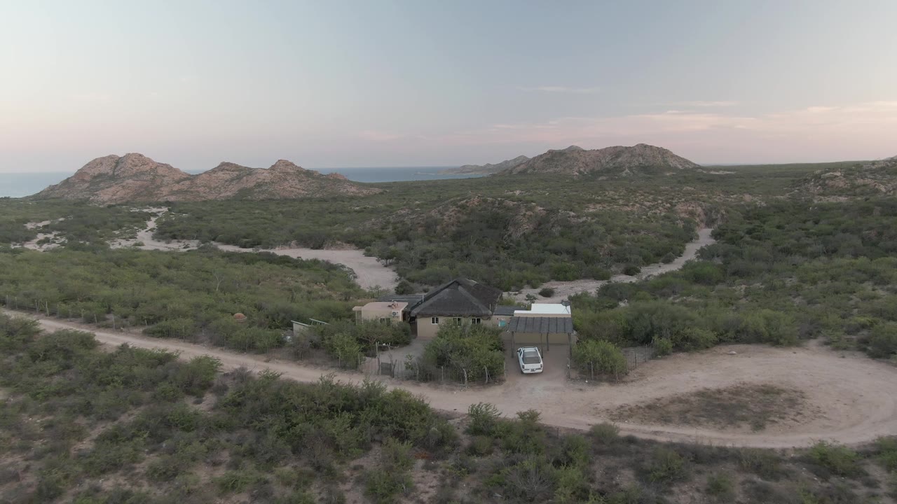 Early aerial view in Cabo Pulmo with the house and car near the lower center, pale dirt tracks crossing the scrub, rocky hills beyond, and no visible people.