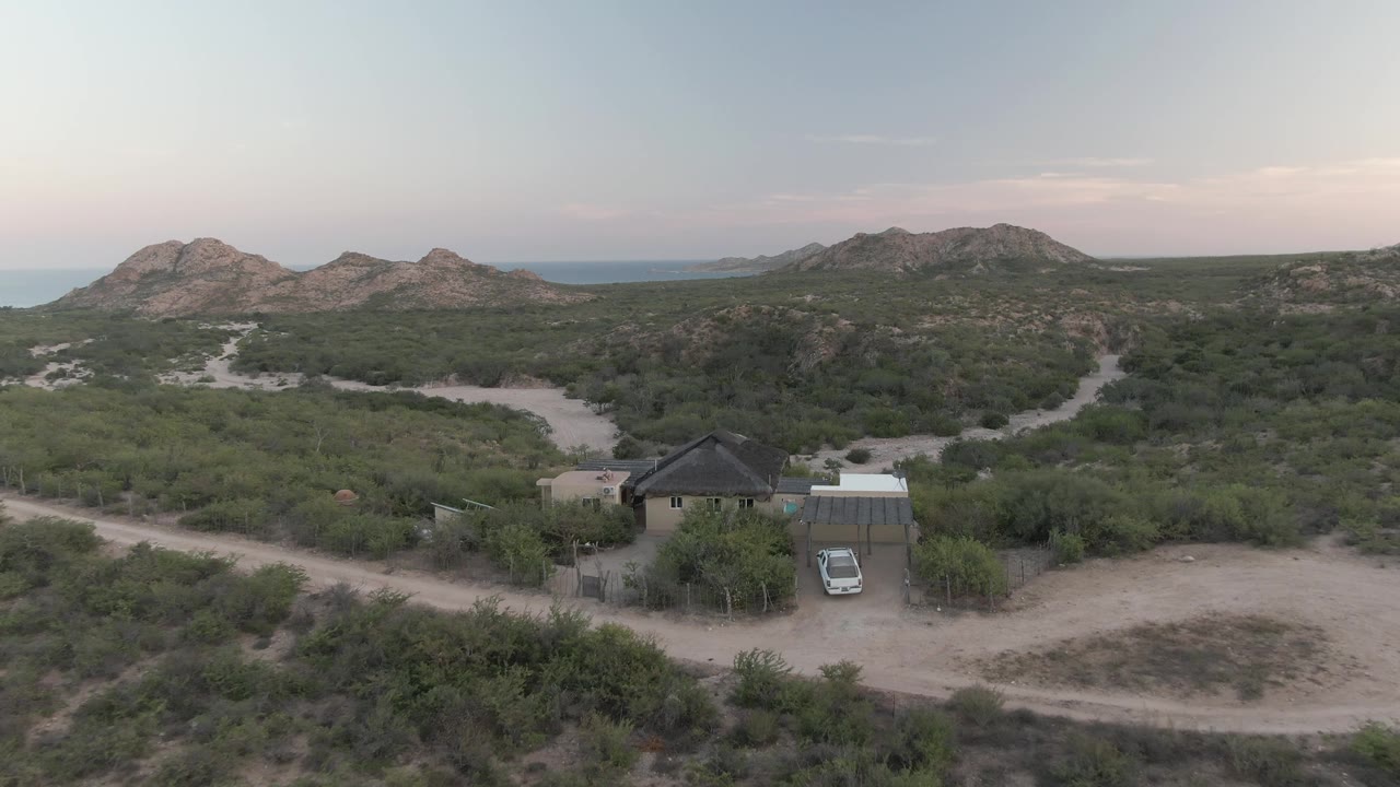 Mid-clip aerial frame over Cabo Pulmo showing the car more clearly in front of the small house, with a curving sandy road, dense low vegetation, mountains, and no visible people.