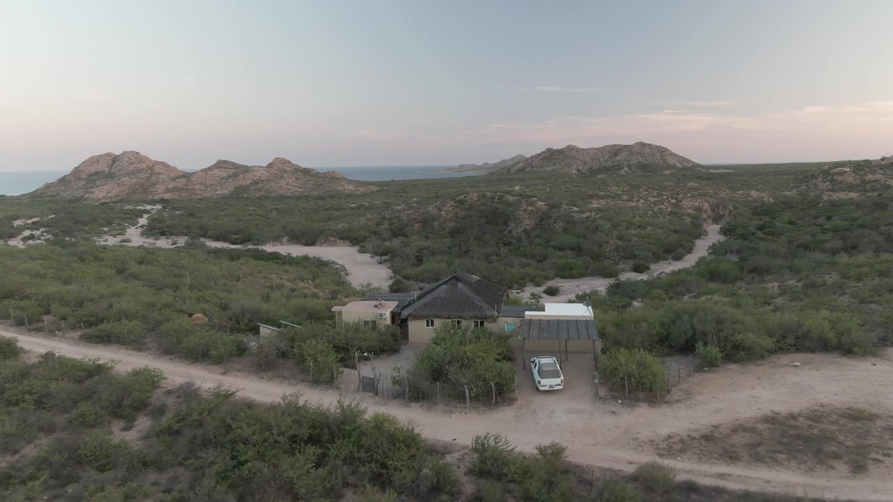 Closer aerial angle on a desert house, parked car, sandy clearing, bushes, and mountain horizon.