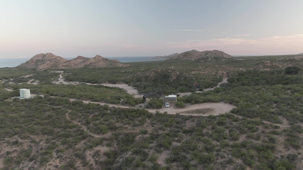 Early aerial frame over Cabo Pulmo desert scrub, showing sandy tracks, a small dark-roofed building, a white tank, a parked white van, rocky hills, a distant sea horizon, and no visible people.