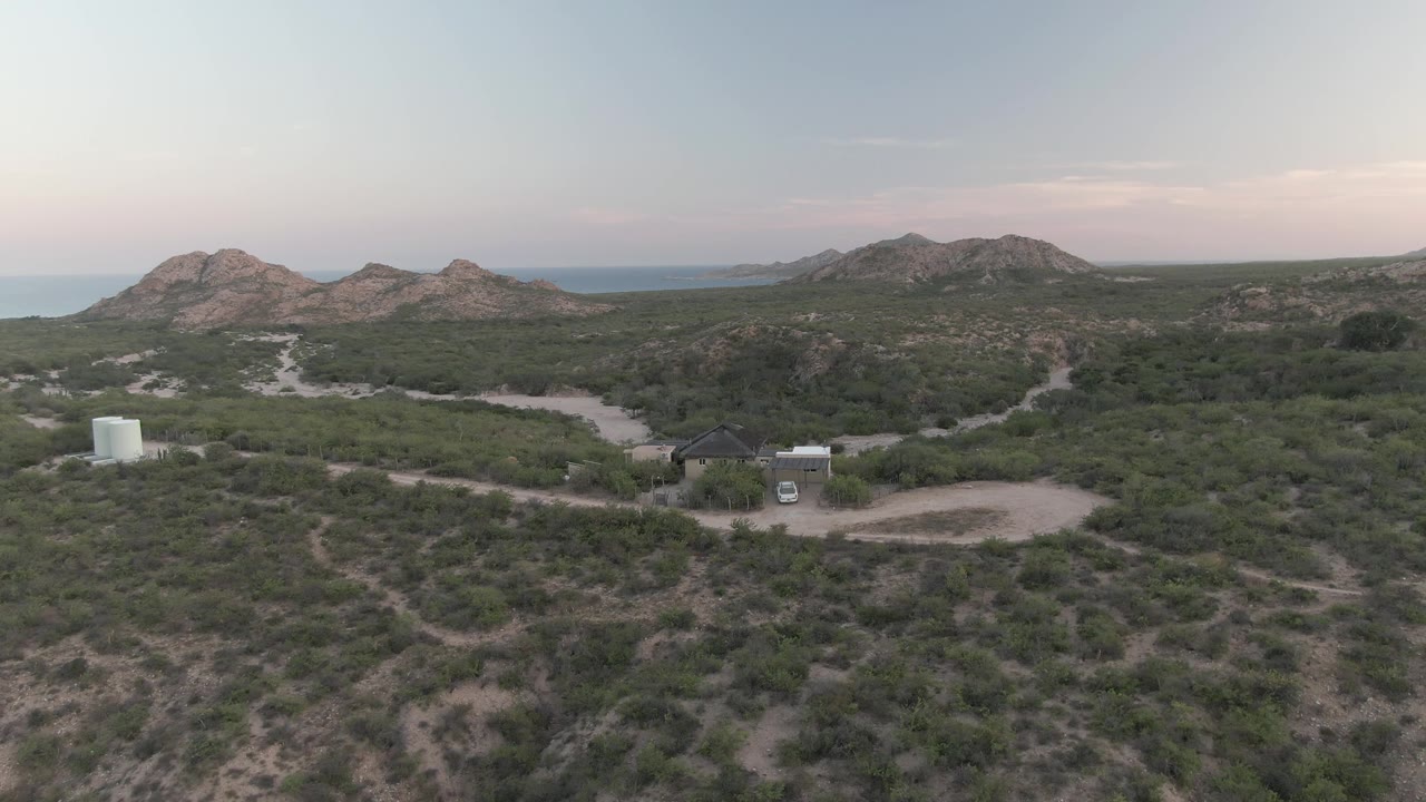 Wide aerial view of scrubland with small building, white tank, dirt road, van, rocky hills, and sea.