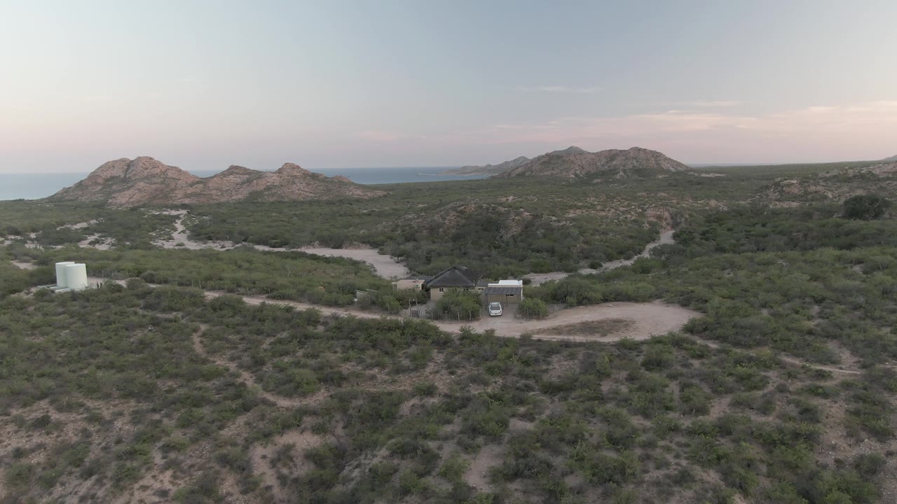 Mid-early frame from the Cabo Pulmo clip with winding dirt tracks crossing green desert brush, a small building and white tank in the midground, the van still near the road, and no visible people.