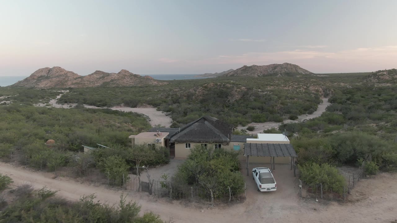 Aerial frame over Cabo Pulmo showing a small desert house, a carport with a white vehicle, pale dirt roads, scrub vegetation, distant low hills, and no visible people.