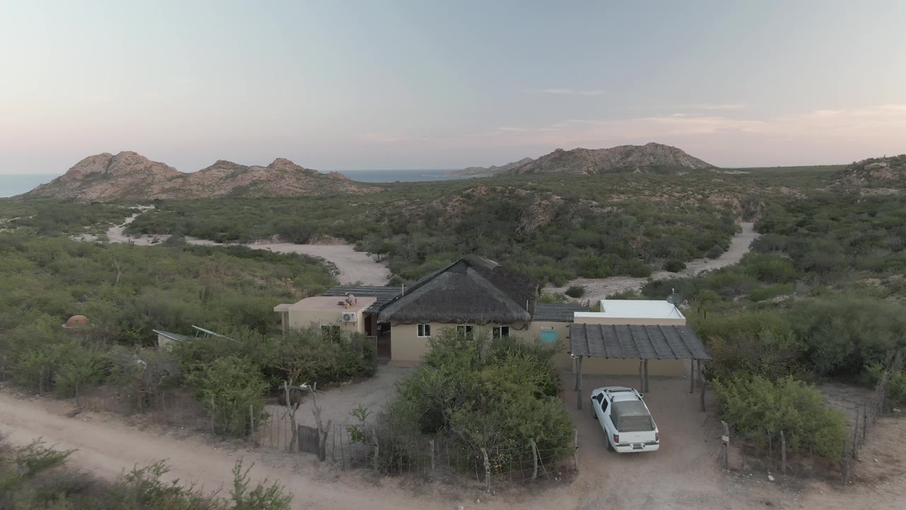 Aerial view in Cabo Pulmo with the rural house and carport slightly closer, a white vehicle below, branching dirt roads through scrub, distant hills, and no visible people.