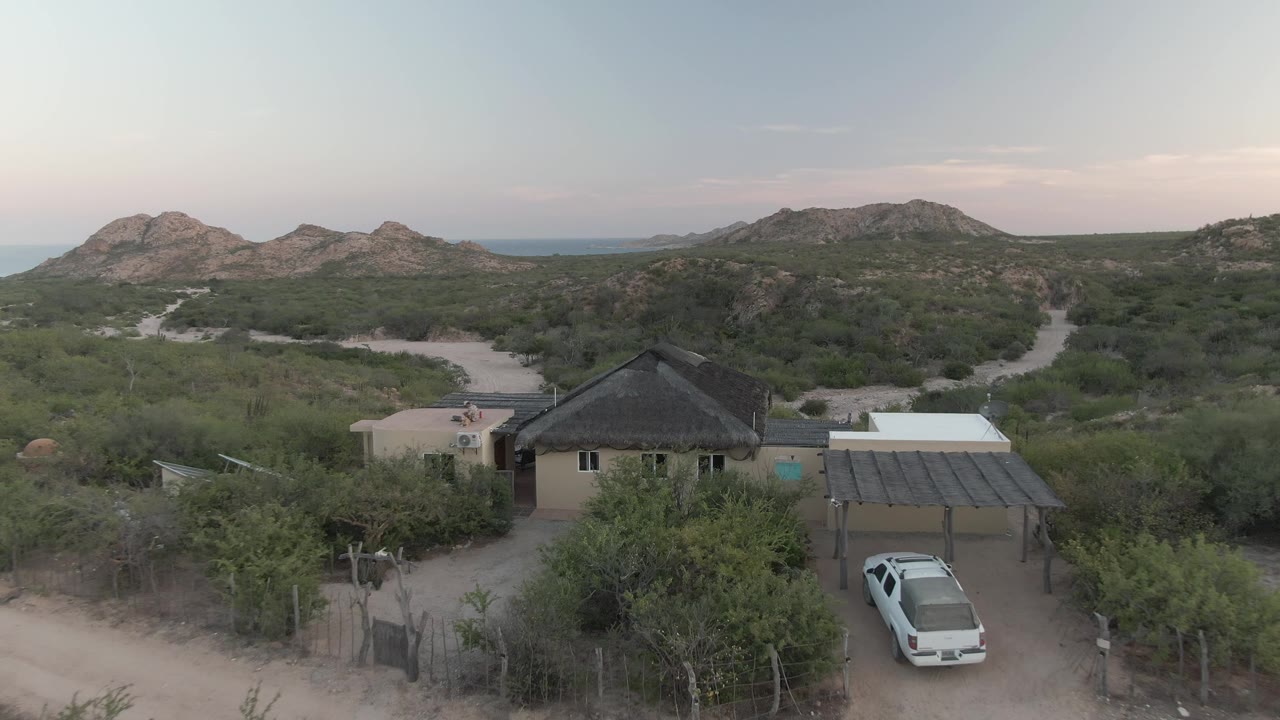 Cabo Pulmo aerial frame showing the house and carport shifted lower in view, with the white vehicle near the driveway, dry green scrub, dirt tracks, low hills, and no visible people.