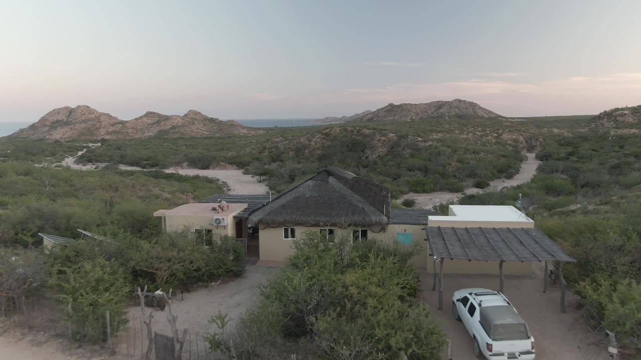 Closer aerial view of a house and carport in desert scrub with a white car below.